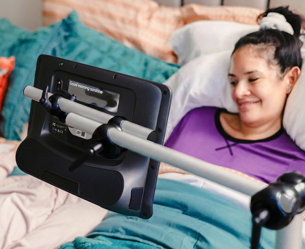Woman with ALS/MND in bed using an AAC device mounted to a floorstand